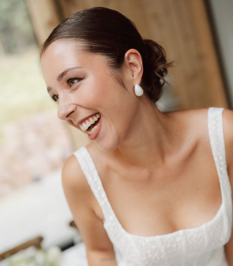 Lächelnde Braut in elegantem Kleid mit Perlenohrringen, vor einem warmen Hintergrund.